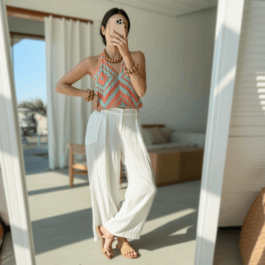 Patterned halter top tucked into white linen pants with wooden bead jewelry and flat sandals for an airy outdoor festive look.
