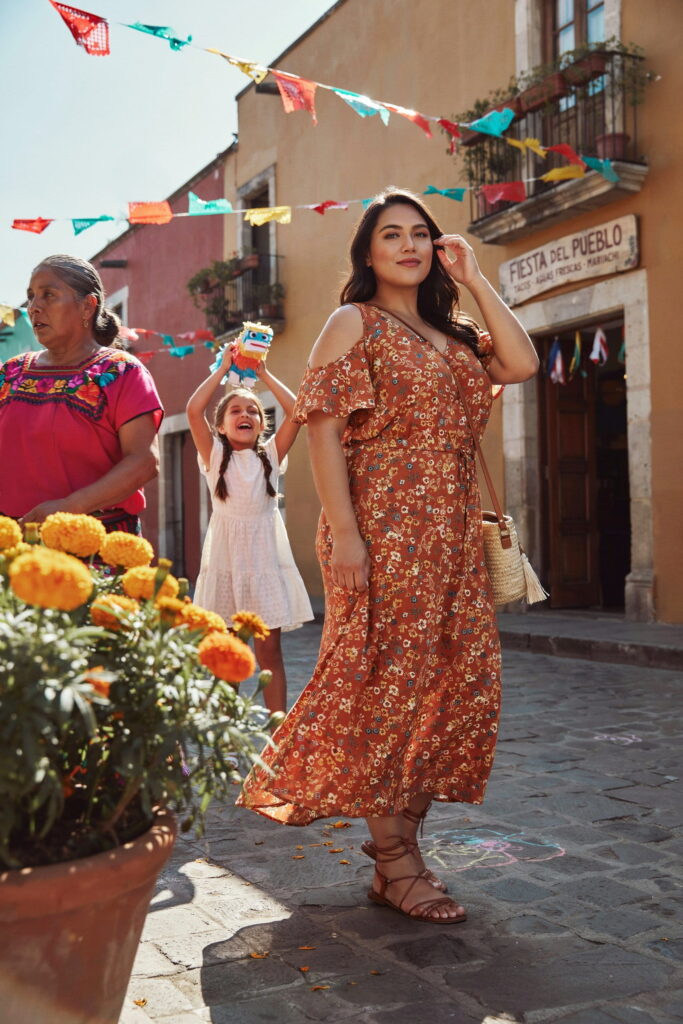 Curvy woman wearing a warm printed cold-shoulder maxi dress with flat strappy sandals at an outdoor Cinco de Mayo street fiesta.
