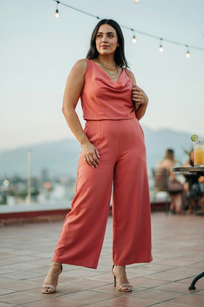 Plus-size woman wearing a deep coral cowl neck wide-leg jumpsuit with nude strappy heeled sandals and layered gold necklaces, creating an elegant and festive Cinco de Mayo evening outfit.