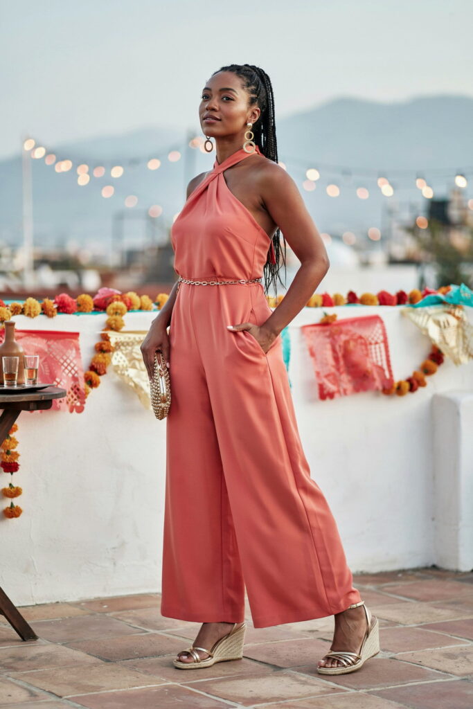 Deep coral wide-leg halter jumpsuit on deep skin styled with stacked gold hoop earrings and gold strappy wedge sandals for an effortless Cinco de Mayo look.