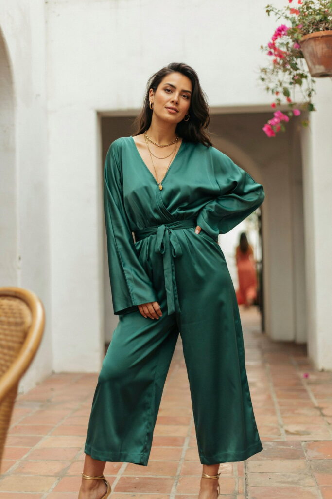 Curvy woman wearing an emerald green deep V-neck wide-leg jumpsuit with a self-tie waist belt, gold strappy heels, and layered gold necklaces for a Cinco de Mayo outfit