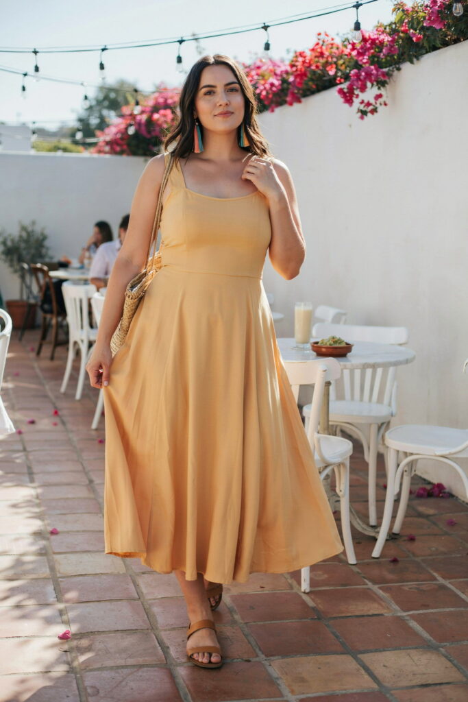 Curvy woman wearing a warm yellow empire waist sundress with tan flat sandals and colorful tassel earrings for a stylish Cinco de Mayo brunch outfit.