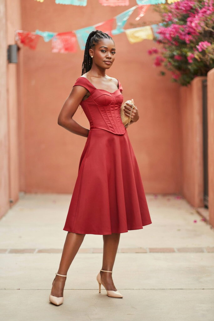 Fire engine red corset top and matching A-line midi skirt on deep brown skin styled with nude pointed-toe kitten heels and small gold studs for a bold Cinco de Mayo outfit.