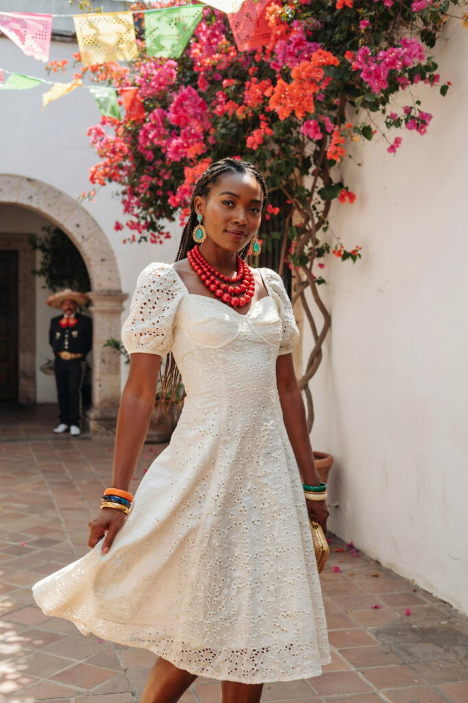 Ivory broderie anglaise knee-length dress on deep brown skin styled with colorful statement jewelry, turquoise earrings, red beads, and stacked bangles for a festive Cinco de Mayo look.