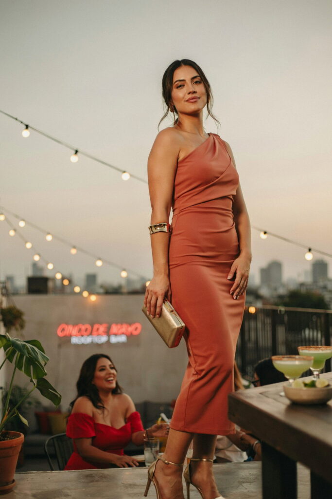 23 Plus Size Cinco de Mayo Outfit Ideas for the Cutest Fiesta Look Ever 21 Woman wearing a terracotta one-shoulder midi dress with gold strappy heels and an oversized gold cuff bracelet, creating a chic and elegant plus-size Cinco de Mayo rooftop bar outfit.